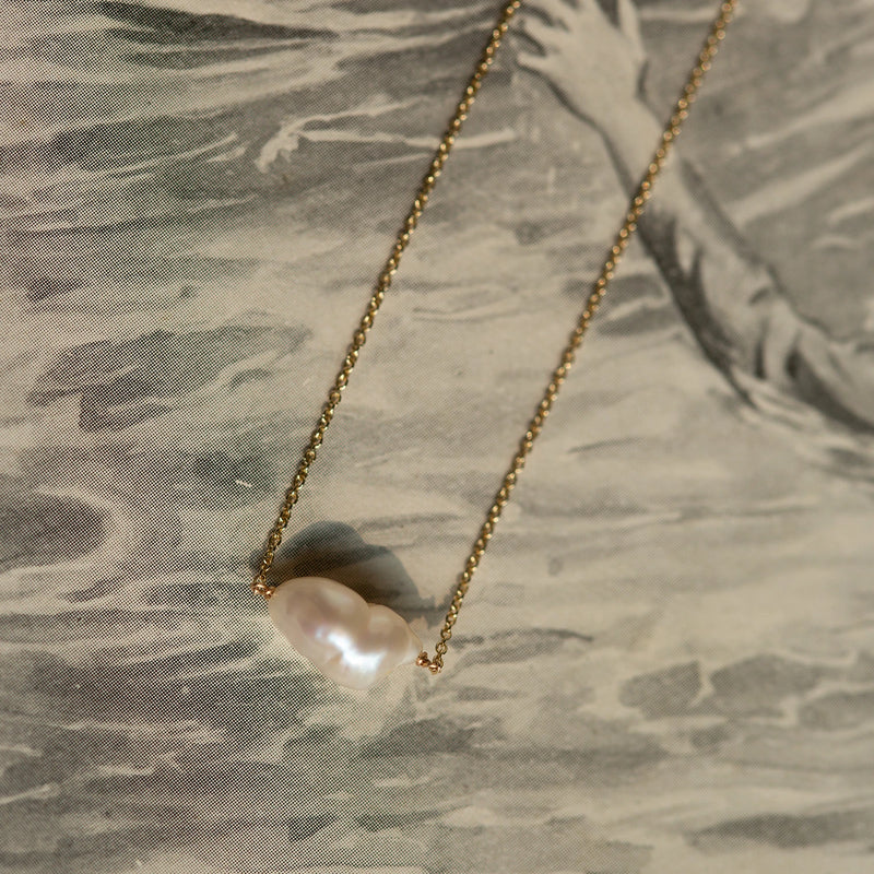 A sofia perla necklace by bluboho, featuring a 14k yellow gold chain with an irregularly shaped pearl pendant, rests on a textured, monochromatic background adorned with a vintage-style illustration of a hand reaching out.