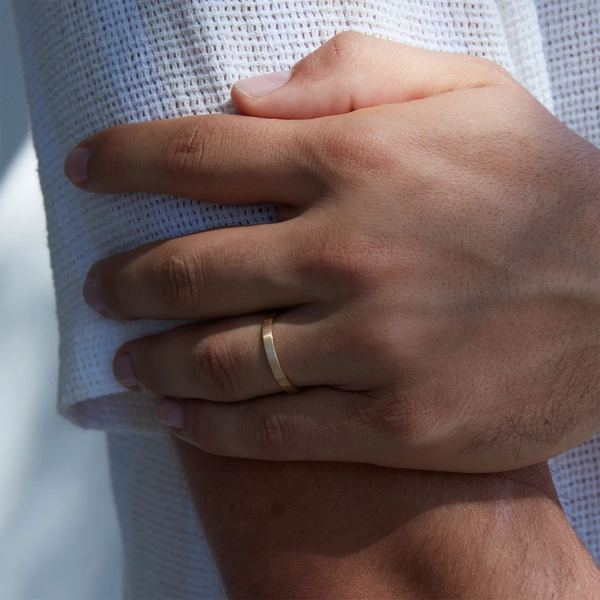 A close-up image captures a hand adorned with the Everlast band brushed - 14k yellow gold from beloved by bluboho, resting on white textured fabric. The arm is crossed, providing a clear view of the understated yet elegant ring. Subtle shadows hint that the photo was taken in natural light.