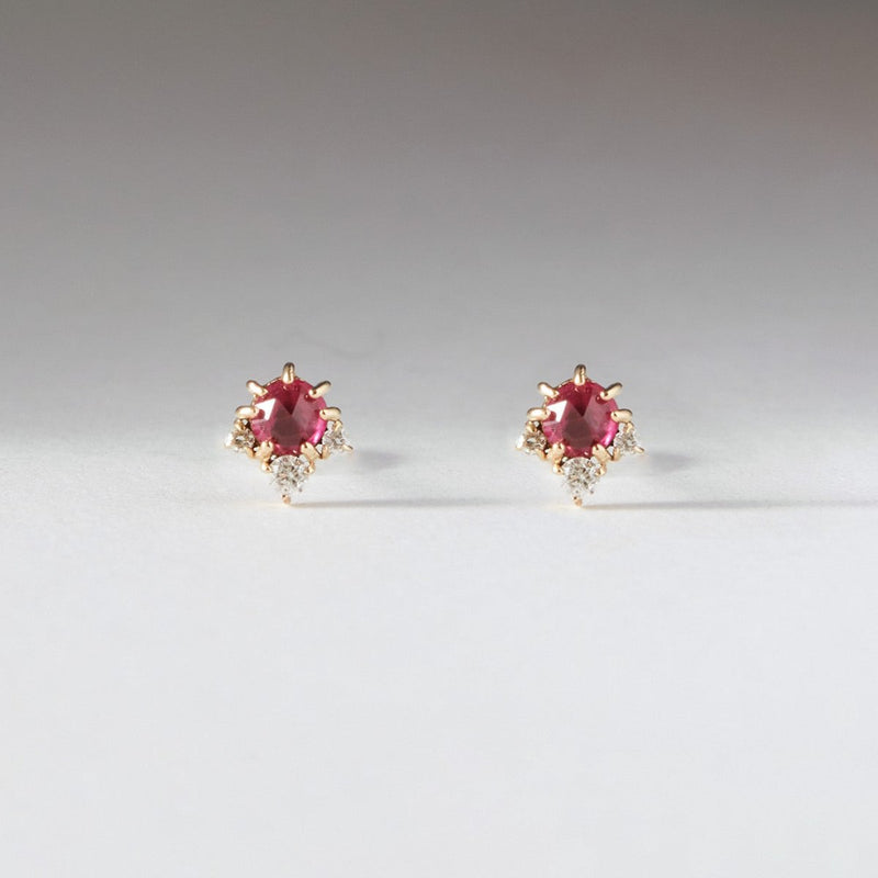 The bluboho full moon glow diamond ruby earrings in 14k yellow gold feature elegant red rubies set in a luxurious gold prong setting. Each earring showcases a small cluster of clear, round diamonds accenting the main ruby gem, and are displayed on a light, neutral background.