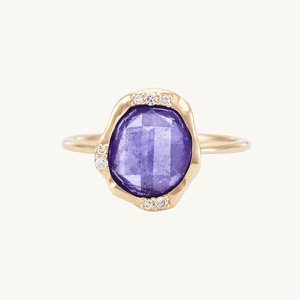 The "tanzanite mood ring" by bluboho features a large, oval-shaped tanzanite gemstone at its center, surrounded by small white diamonds set into the 14k yellow gold band in a wavy, organic design. This ring has a delicate and elegant appearance with a smooth finish.