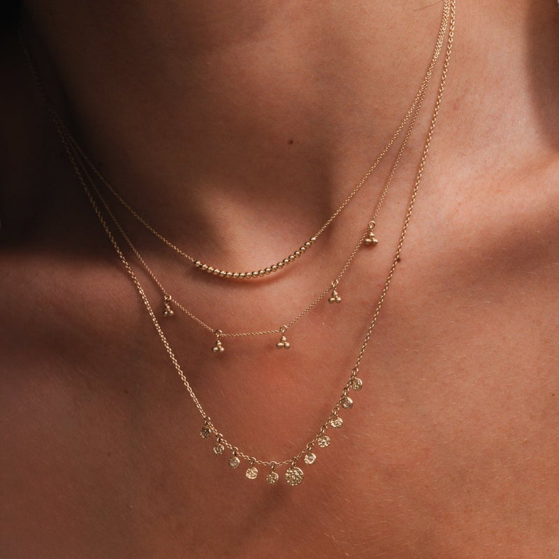 Close-up of a person wearing three layered gold necklaces from bluboho. The top abacus tripod necklace features small beads, the middle one showcases tiny triangular pendants, and the bottom necklace has evenly spaced circular charms. The background is the person’s bare neck and collarbone.