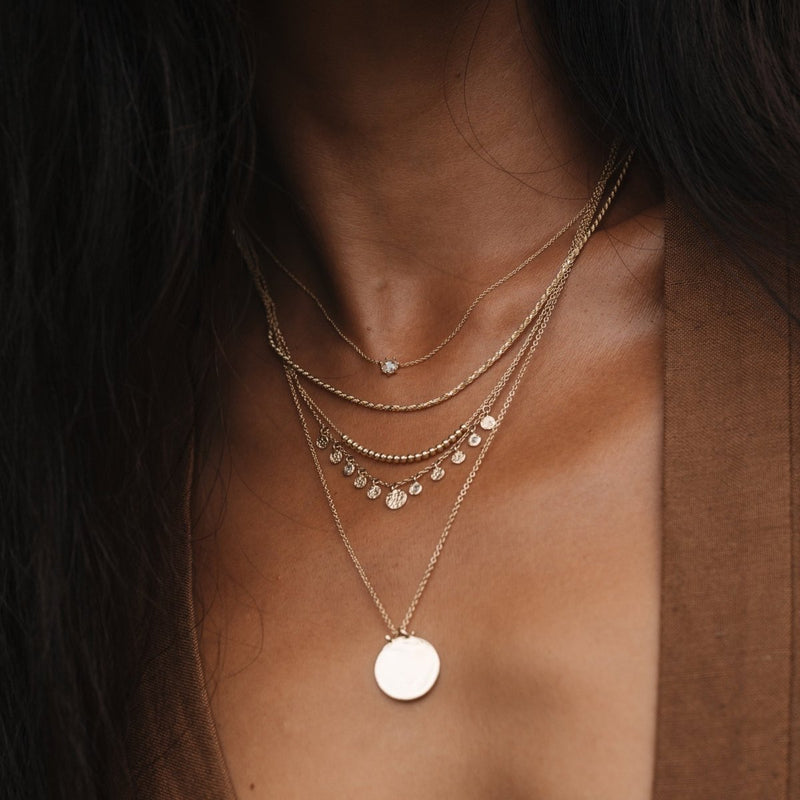 A close-up of a person wearing multiple layered gold necklaces from bluboho, including the abacus disk diamond necklace in 14k yellow gold, is shown. The necklaces feature different designs: a small diamond pendant, a strand adorned with tiny discs, and a larger round pendant. The individual is dressed in a brown top with their hair cascading down on both sides.