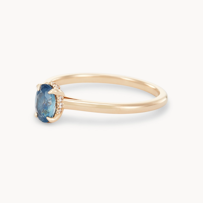 The astral love one-of-a-kind 14k yellow gold ring from beloved by bluboho features a single oval-shaped blue sapphire centerpiece surrounded by small diamond accents. The thin gold band is simple and elegant, perfectly complementing the ornate setting of the gemstone.