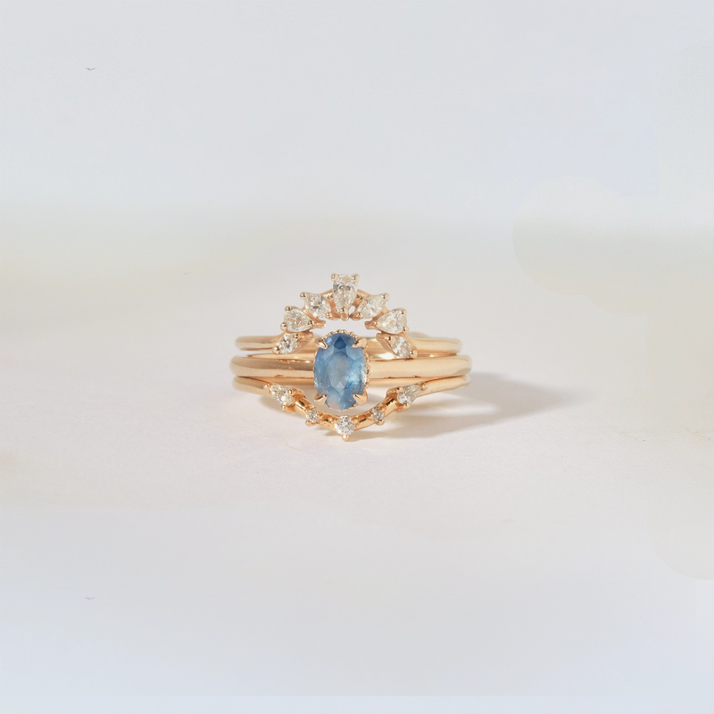 The astral love one-of-a-kind 14k yellow gold ring from beloved by bluboho, featuring a central blue sapphire and multiple smaller diamonds in an elegant design, is displayed against a soft white background. The intricate detailing of the ring enhances its luxurious and sophisticated appearance.