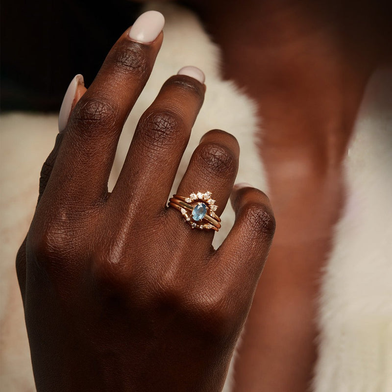 A close-up image of a hand showcasing elegant jewelry. The hand is adorned with a stack of gold rings, featuring the "astral love one-of-a-kind - 14k yellow gold ring, blue sapphire" by beloved by bluboho. The prominent blue gemstone is surrounded by smaller white stones. The nails are manicured with light-colored polish, and the background is softly blurred.