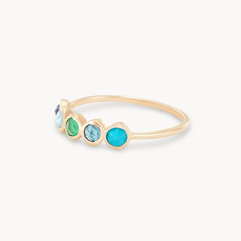 The Coastal Nomad Five Stone Mood Ring by bluboho is a delicate 14k yellow gold ring featuring five round gemstones set in a row. The stones include tanzanite, larimar, emerald, London blue topaz, and turquoise, creating a vibrant and elegant array of shades in blue and green. The thin and simple band draws attention to the colorful stones.