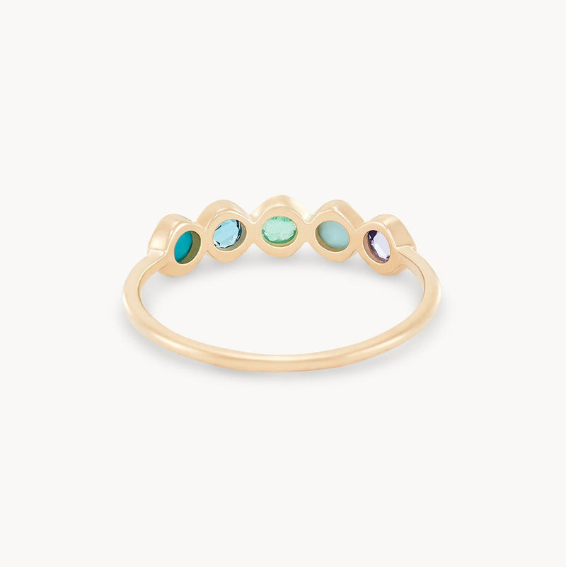 The Coastal Nomad Mood Ring by bluboho is a delicate piece crafted in 14k yellow gold, featuring tanzanite, larimar, emerald, London blue topaz, and turquoise gemstones set in an open bezel setting. The smooth and polished band complements the varied colors of green, blue, and purple stones for a simple yet elegant appearance.

