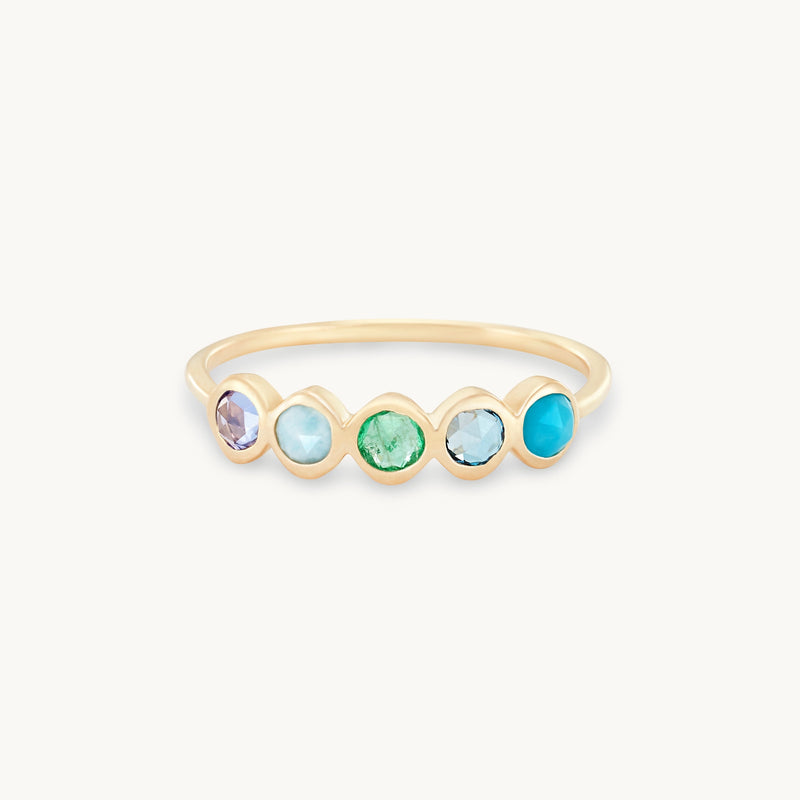 The Coastal Nomad five stone mood ring by bluboho is a delicate 14k yellow gold ring set with five evenly spaced gemstones. The gemstones are tanzanite, larimar, emerald, London blue topaz, and turquoise, each in a round bezel setting that creates a colorful and elegant design against the plain white background.