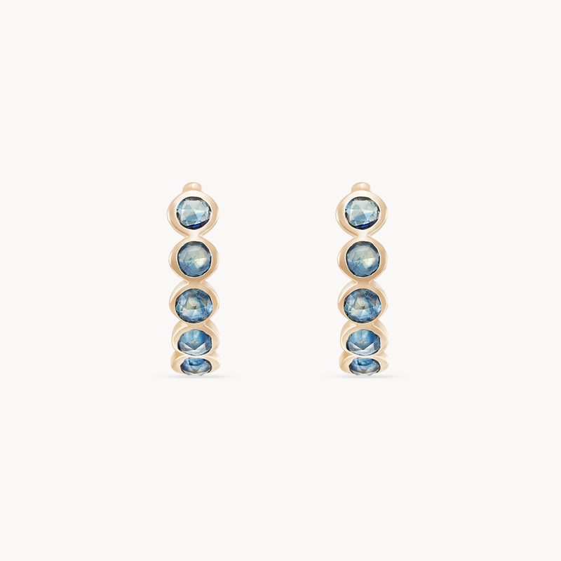 Introducing the bluboho blue sapphire five stone mood hoops, crafted in 14k yellow gold and adorned with a vertical row of five round blue sapphires. These elegant earrings are beautifully showcased against a plain white background.