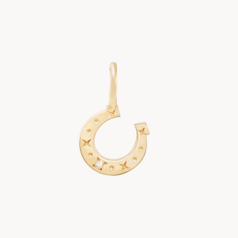 Close-up of the bluboho Wild Luck Horseshoe Charm - 10k Yellow Gold, Diamond. The charm is shaped like a horseshoe and adorned with small embossed star shapes and tiny round studs. One of the star shapes holds a small sparkling diamond, while the attached hoop remains simple and unadorned.