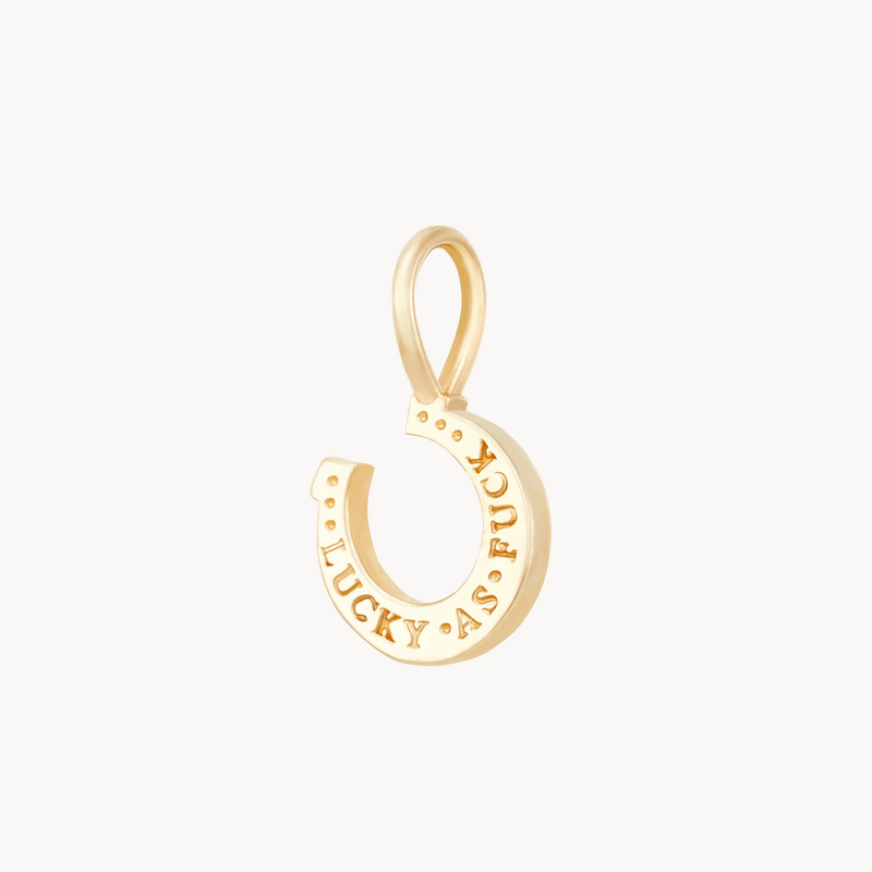 The Wild Luck Horseshoe Charm by bluboho is crafted from 10k yellow gold and inscribed with the words "LUCKY AS FUCK." This pendant features a smooth, polished finish and includes a small loop at the top for easy attachment to a chain or bracelet.