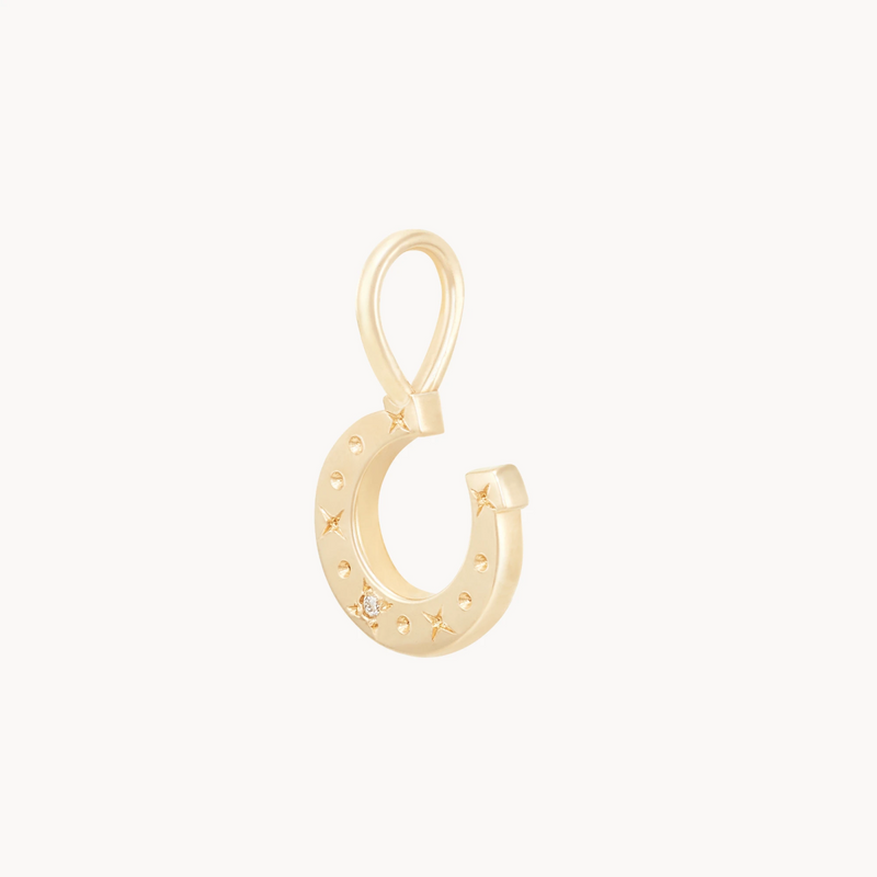 The wild luck horseshoe charm by bluboho is crafted from 10k yellow gold and adorned with small star engravings, featuring a tiny diamond near the bottom. It includes a loop at the top for attaching to a necklace or bracelet, all set against a white background.