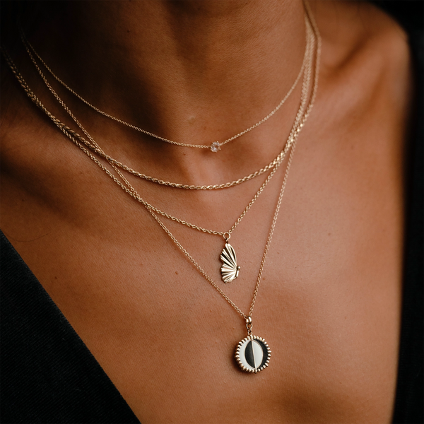 A close-up image of a person's neck adorned with four delicate gold necklaces, including the Metamorphosis Butterfly Wing Necklace by bluboho. The layered look features a small chain with a tiny star pendant, another chain showcasing the butterfly wing-shaped pendant in 14k yellow gold, and an additional chain with a round pendant embedded with either pearl or stone.