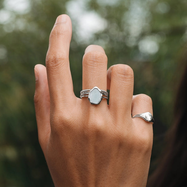 Close-up of a hand adorned with two rings: the middle finger features the blue chalcedony mood ring from bluboho, showcasing a hexagonal, pale blue stone set in sterling silver, while the pinky finger sports a simple, sleek silver band. The background is blurred with green foliage.