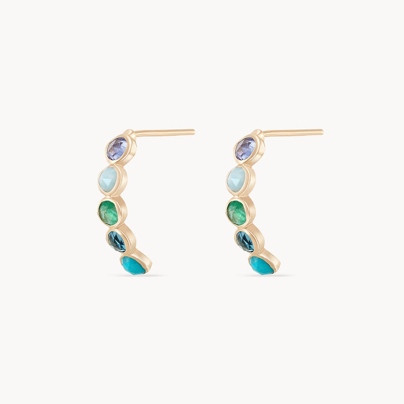 A pair of bluboho coastal nomad five stone mood hoops - 14k yellow gold, featuring tanzanite, larimar, emerald, london blue topaz, and turquoise. The earrings have a modern design with a subtle curve, presenting a bold yet elegant aesthetic. The background is plain white.