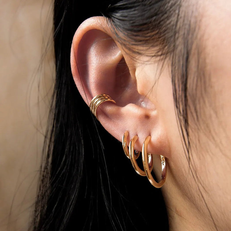 Close-up of a person's ear adorned with multiple gold hoop earrings. The ear features a single gold cuff at the top cartilage area and three progressively larger bluboho dagger medium hoop - 14k yellow gold hoops through the earlobe. Dark hair partially frames the ear.