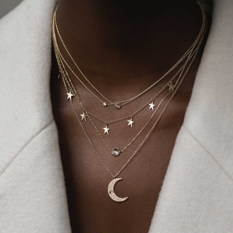 A person adorned with the extra large supernova necklace in 14k yellow gold from bluboho, showcasing a crescent moon pendant, star charms, and delicate chains with small beads, elegantly styled over a pale, textured garment.