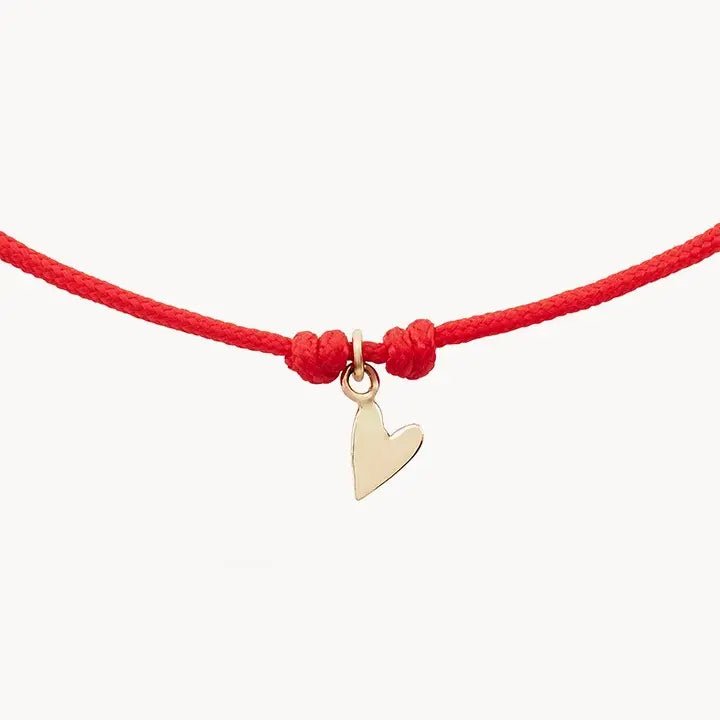 The Deep Love Contemplation Cord Bracelet by bluboho features a small 10k yellow gold heart charm elegantly positioned at the center of a thin red cord. The charm is securely fastened between two knots, resulting in a simple and stylish accessory. The bracelet is showcased on a plain white background.