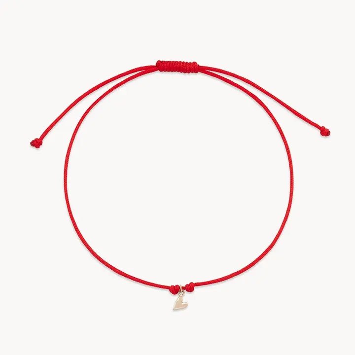 The deep love contemplation cord bracelet from bluboho features a minimalist red string with adjustable knots on both ends and a small, 10k yellow gold heart-shaped pendant. The bracelet is elegantly displayed against a plain white background.