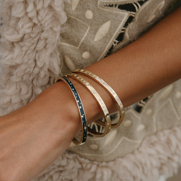 A close-up of a person's arm adorned with four bluboho bangles. One bangle is the dream state bangle in 14k yellow gold with blue enamel and small gold stars, while the other three are plain gold. The person is wearing a cream-colored textured fabric with embroidered details.