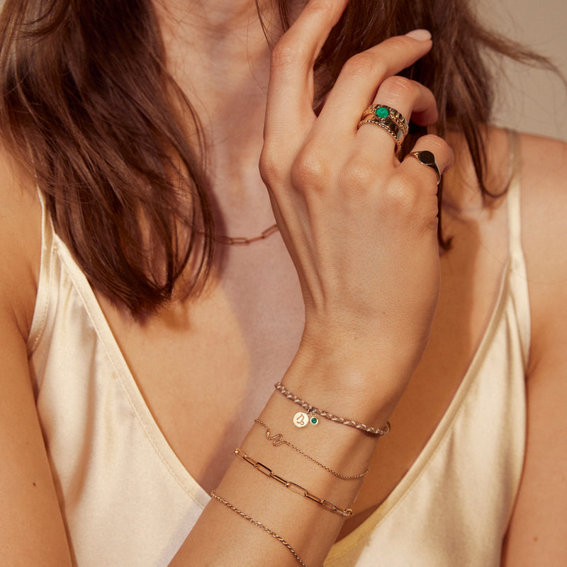 A woman wearing a beige satin top showcases her jewelry. She wears several gold bracelets, a necklace, and multiple rings, including one with a turquoise stone and another with an oval black stone. On her wrist is the virgo zodiac earth element cord bracelet - 10k yellow gold, emerald, cord by bluboho. Her fingers touch her face, highlighting the accessories.