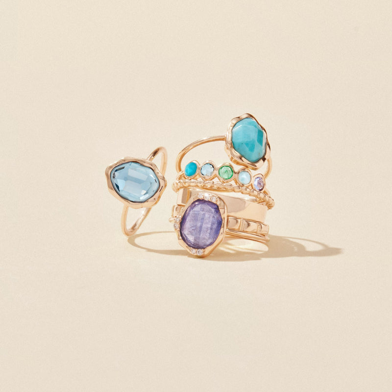 A collection of five Coastal Nomad rings by bluboho, set in 14k yellow gold and featuring tanzanite, larimar, emerald, London blue topaz, and turquoise stones. Each ring showcases unique designs, from simple single-set stones to intricate multi-stone arrangements, all displayed artfully on a pale beige background.