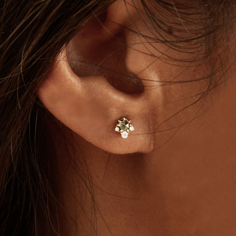 Close-up image of an ear wearing bluboho's Full Moon Glow Diamond London Blue Topaz Earring, featuring a small blue topaz gemstone with a diamond beneath it, set in 14k yellow gold. The background is an out-of-focus representation of hair and skin tone.