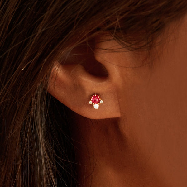 Close-up of a person's ear wearing a stud earring. The earring, identified as the Full Moon Glow Diamond Ruby Earring by Bluboho, features a vibrant ruby gemstone surrounded by small, sparkling diamonds, set in 14k yellow gold. The person has long, straight, dark hair.