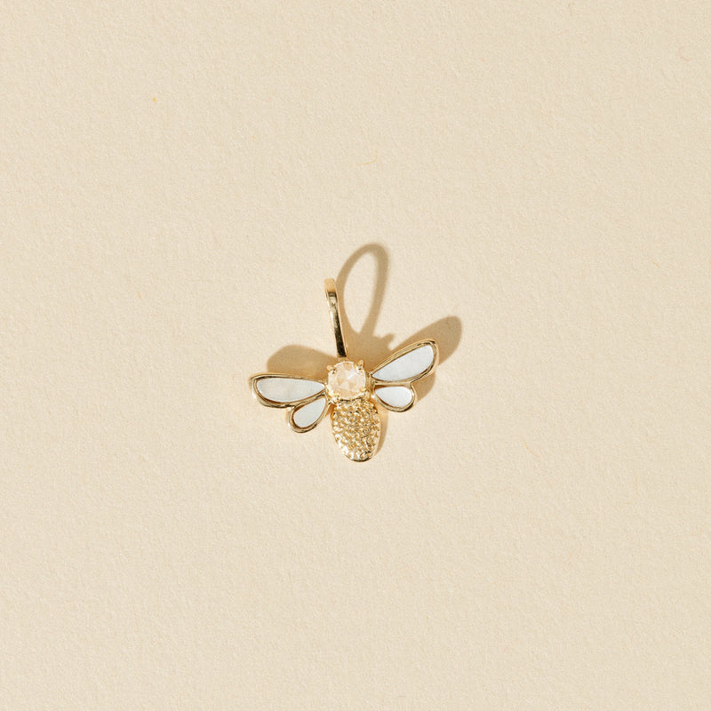 The honey glow bee charm by bluboho, crafted from 10k yellow gold and featuring a mother of pearl inlay, lies on a beige textured background. The charm is finely detailed and has a loop at the top for threading.