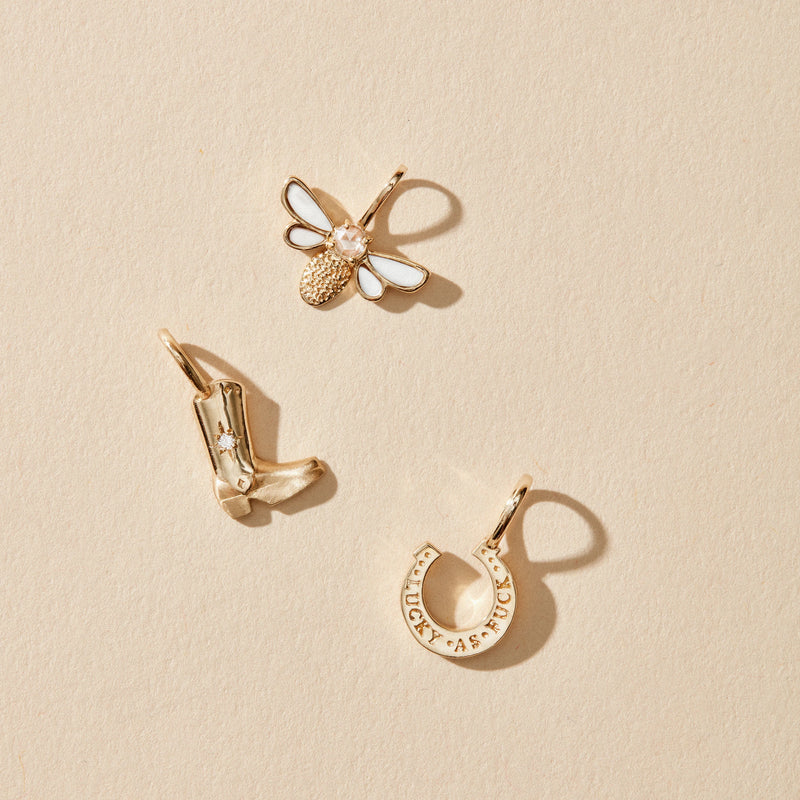 Three small gold charms are displayed on a beige surface. The charms include a honeybee with white wings and a textured body, a cowboy boot adorned with a small star, and the wild luck horseshoe charm by bluboho in 10k yellow gold and diamond, inscribed with the word "lucky" around its outer edge.