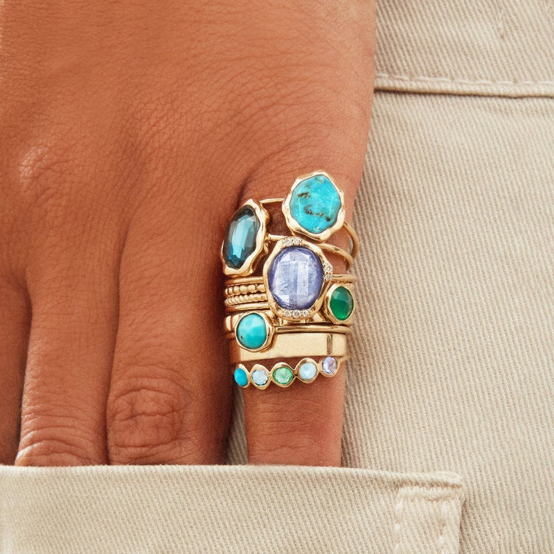 A hand adorned with the Coastal Nomad Five Stone Mood Ring by bluboho, featuring 14k yellow gold band set with tanzanite, larimar, emerald, London blue topaz, and turquoise stones, is tucked into the pocket of beige pants. The rings vary in design, creating a layered and eclectic look.