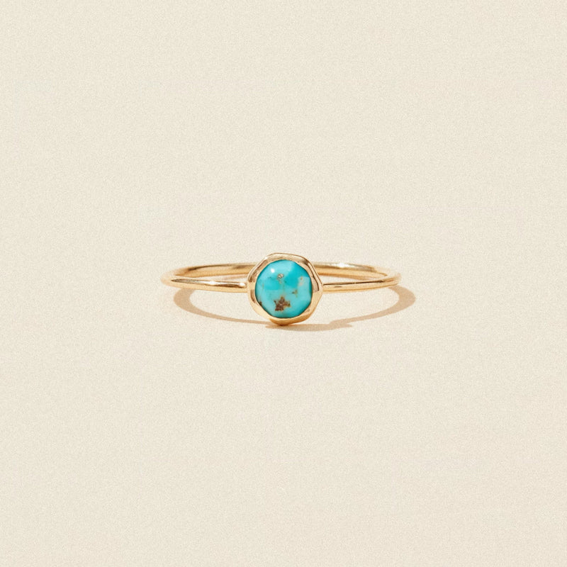 A delicate gold ring with a thin band, featuring a small round turquoise gemstone set in the center. The turquoise stone showcases natural variations in color, with a mix of blue and brown hues. The ring is displayed against a simple beige background. This is the bluboho turquoise mini mood ring - 10k yellow gold, turquoise.