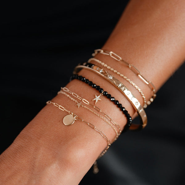 A close-up of a wrist adorned with multiple gold and black bracelets of varying designs, including the bluboho guidance bright star black spinel stretch bracelet - 14k yellow gold. The collection features chains with charms, a black bead bracelet, and bangles with star motifs and textured finishes, elegantly layered on a person's skin.