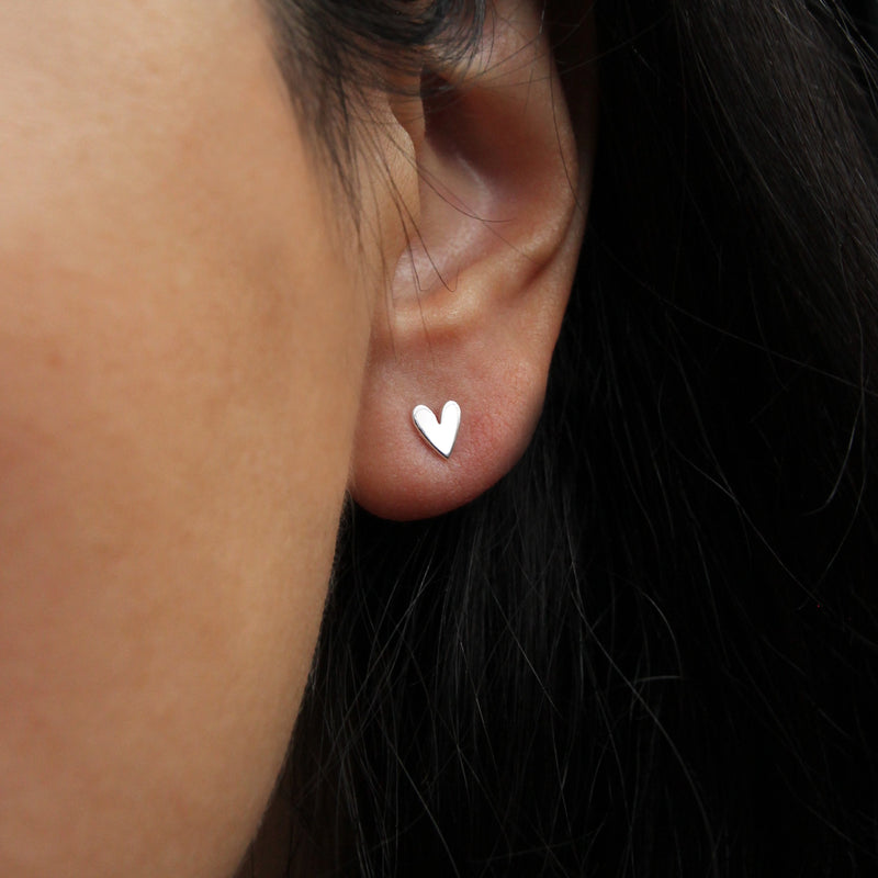 Close-up of a person's ear wearing a small lovely heart earring silver - sterling silver by bluboho. The background shows dark hair framing the ear.