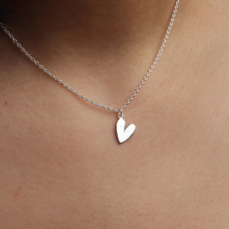 A close-up of a person wearing the bluboho sterling silver "Lovely Necklace Silver" with a small, heart-shaped pendant. The pendant is smooth and polished, hanging from a delicate silver chain that rests gracefully against the person's skin. The neutral background keeps the focus squarely on this exquisite necklace.