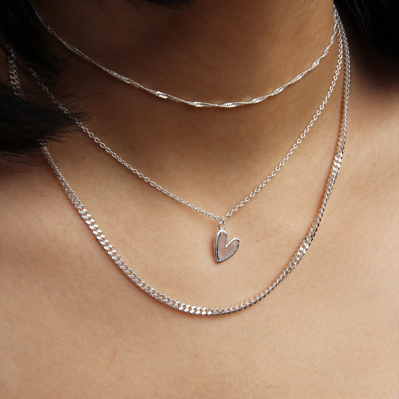  Close-up photo featuring three "lovely necklace silver" pieces by bluboho on a person's neck. The top piece is a delicate sterling silver chain, the middle one is a sterling silver chain adorned with a heart-shaped pendant, and the bottom piece is a thicker, flat sterling silver chain. The backdrop showcases the person's skin with a glimpse of dark hair visible in the frame.