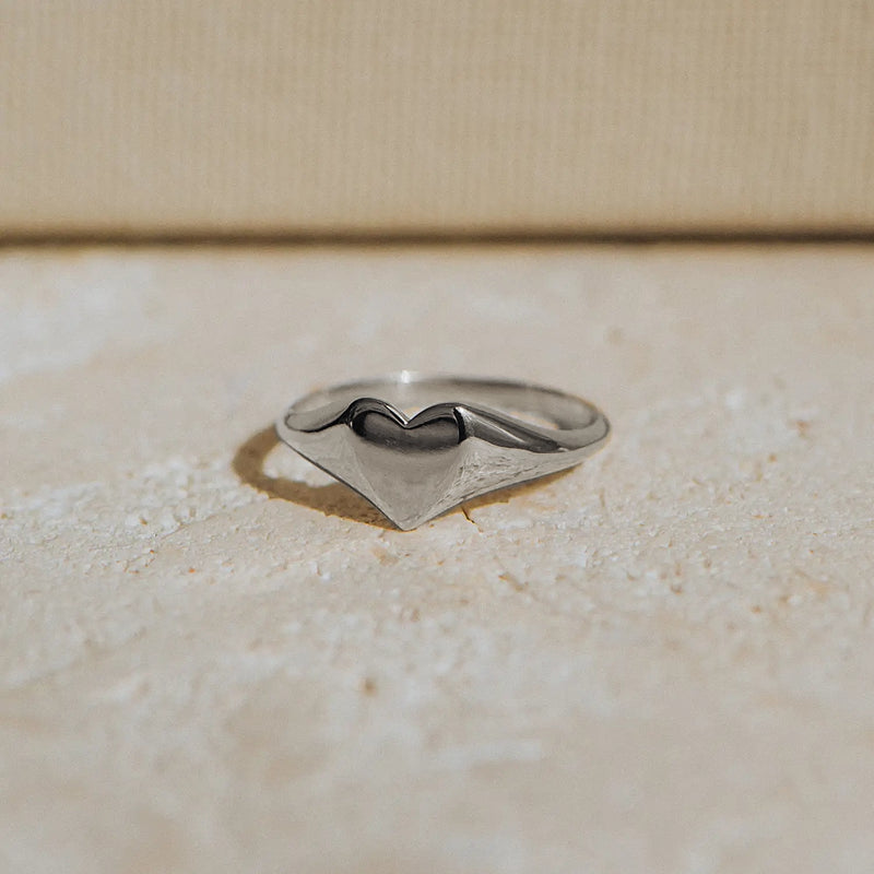 A lovely heart signet ring from bluboho, crafted in sterling silver, lies on a textured beige surface. The ring boasts a minimalist design with a singular, prominent chevron shape creating a gentle peak at the center. The background is softly lit, enhancing the ring's polished and reflective surface.