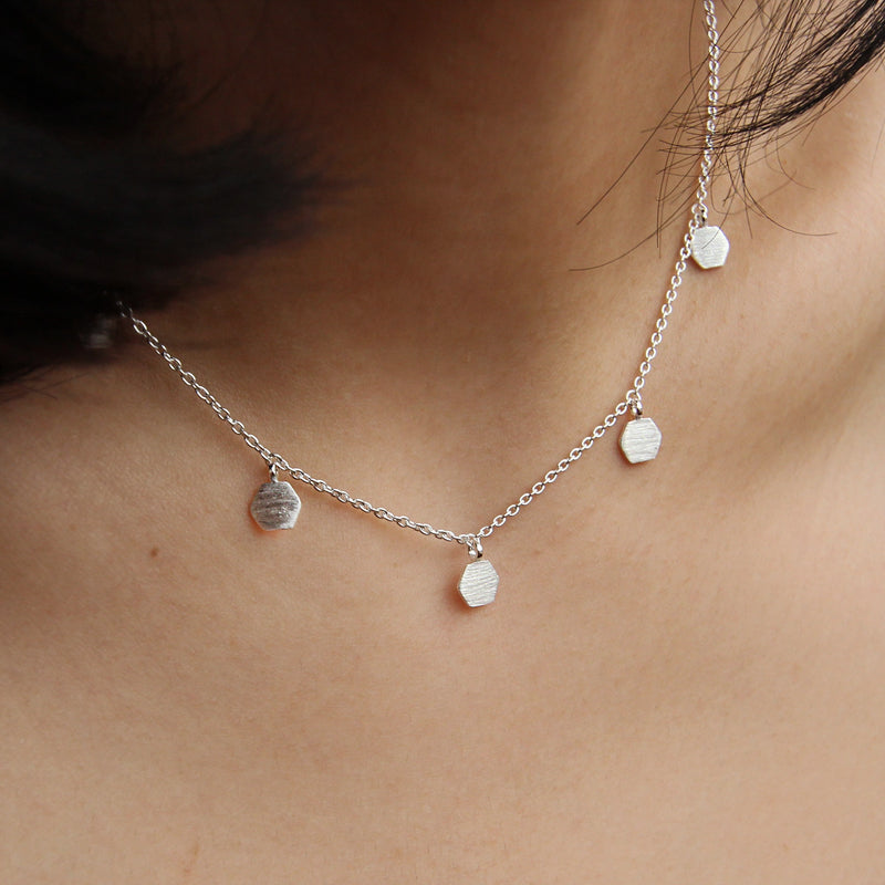 A close-up shot features the honeysuckle necklace in silver by bluboho, crafted from sterling silver. The delicate chain showcases small hexagonal pendants evenly spaced along its length, resting gracefully against the neck's skin with strands of dark hair partially visible in the background.