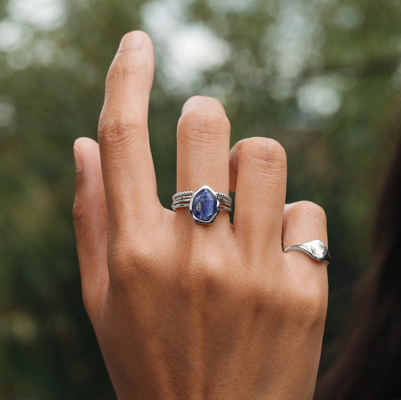 A close-up of a hand showcasing two rings. The middle finger is adorned with the bluboho lapis lazuli mood ring in sterling silver, featuring a large, blue, hexagonal gemstone. The pinky finger sports a simple silver band. The background reveals blurred greenery.