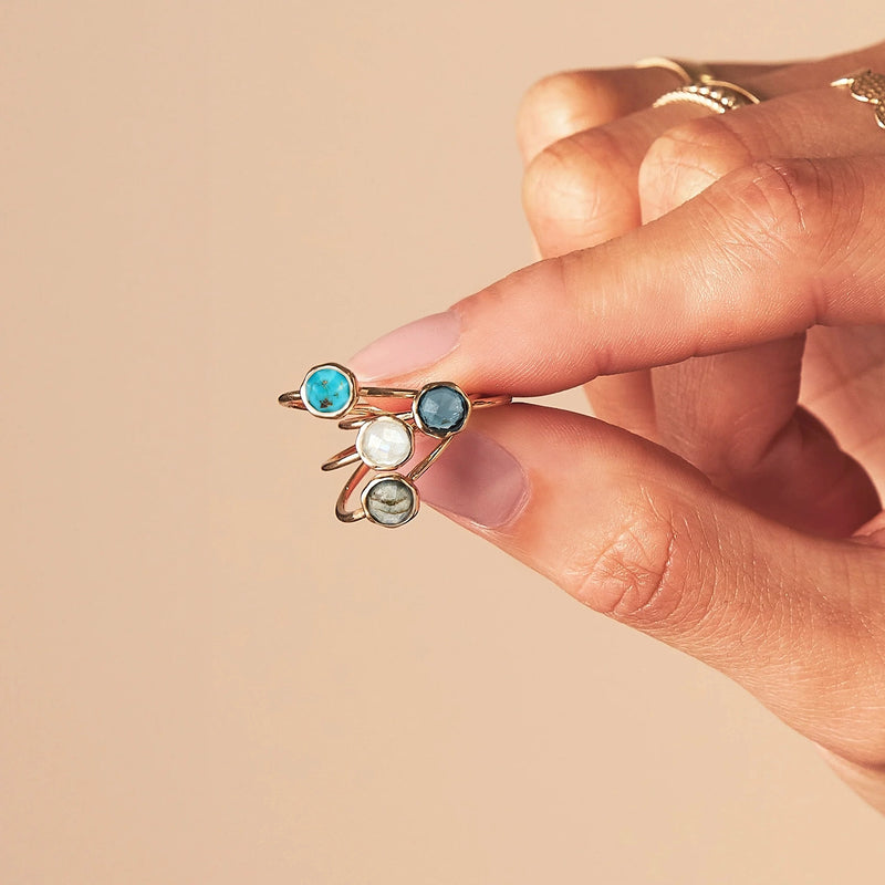 A hand holding three delicate gold rings, each adorned with a different colored gemstone: turquoise blue, deep blue, and clear. The background is a muted beige. The hand is also wearing the bluboho turquoise mini mood ring in 10k yellow gold on the index finger.