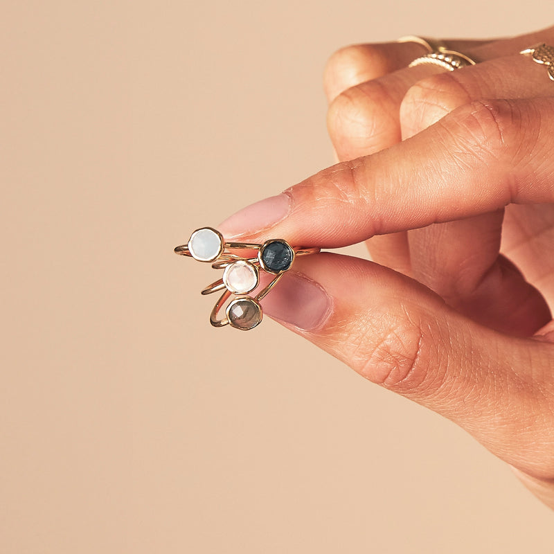 A hand with minimalistic gold rings on its fingers holds a stack of four delicate gold rings, each featuring a different gemstone: a white stone, a black stone, a clear stone, and an iridescent stone. The background is a soft beige color. The rings are the moonstone mini mood ring - 10k yellow gold, moonstone from bluboho.