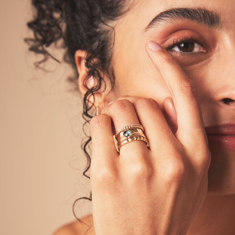 A close-up image of a person touching their face with their fingers, showcasing three stunning london blue topaz mini mood rings by bluboho, crafted in 10k yellow gold and adorned with faceted blue topaz gemstones on their index finger. The person has curly hair, smiles gently, and stands against a neutral beige background.