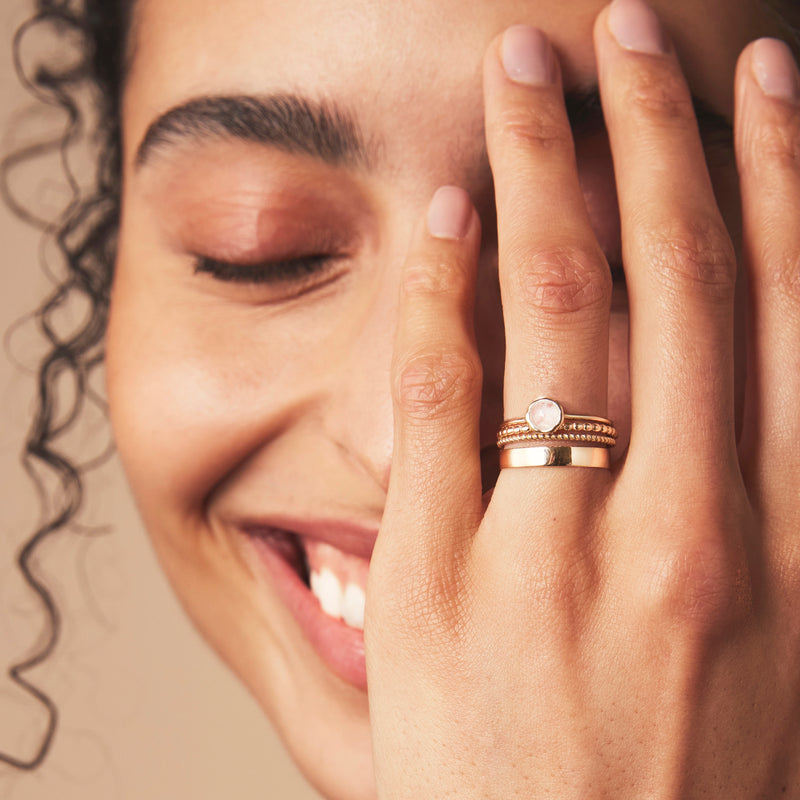 A person with curly hair smiles gently while covering one eye with their hand, which showcases multiple gold rings, including the bluboho moonstone mini mood ring - 10k yellow gold, moonstone. The manicure is clean and simple, with nails painted in a soft, neutral color. The background is beige.