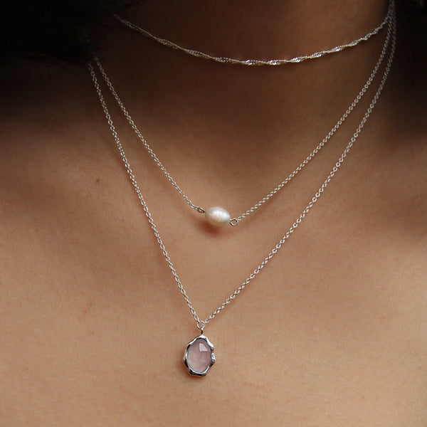 A close-up image of a person's neck showcasing three layered necklaces. The top necklace features a thin, simple chain, the middle one boasts a small white pearl, and the bottom piece is the "blue chalcedony mood necklace" from bluboho, highlighting an oval-shaped blue chalcedony stone framed in sterling silver.