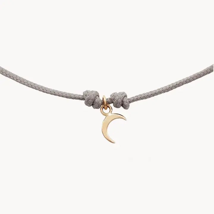 The moon phases contemplation cord bracelet by bluboho showcases a small, 10k yellow gold crescent moon charm at its center, adorning a light grey cord tied with simple knots on either side. The backdrop is a solid white.