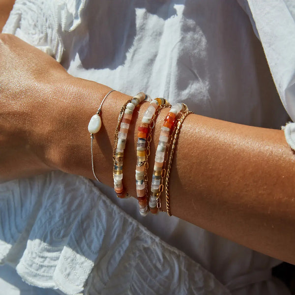 A close-up of a person's arm showcases a beautiful array of bracelets. Among them is the sand dune bead bracelet by bluboho, featuring a beige cord adorned with gemstone beads. The collection also includes a delicate thin chain bracelet, a pearl-accented bracelet, and several beaded bracelets in white, gold, and red tones. The person is elegantly dressed in white ruffled fabric.