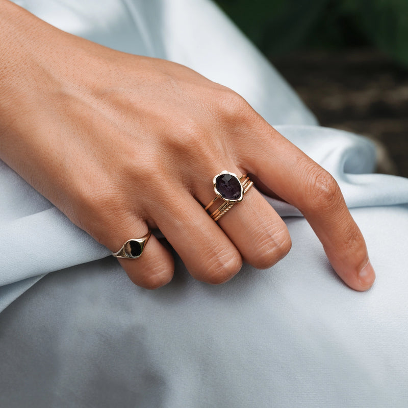 A close-up of a hand adorned with two rings: one gold ring featuring a purple gemstone on the middle finger, and another bluboho black onyx mood ring in 14k yellow gold on the pinky finger. The hand rests gently on a light blue satin fabric.