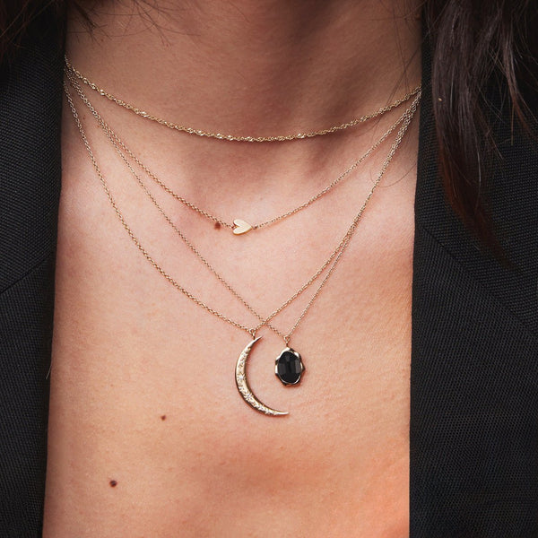 Close-up of a person's neck adorned with four layered necklaces. The top necklace has a small heart pendant, the second one is plain, the third features a crescent moon with small stones, and the fourth is the black onyx mood necklace from bluboho. The person wears a dark top.