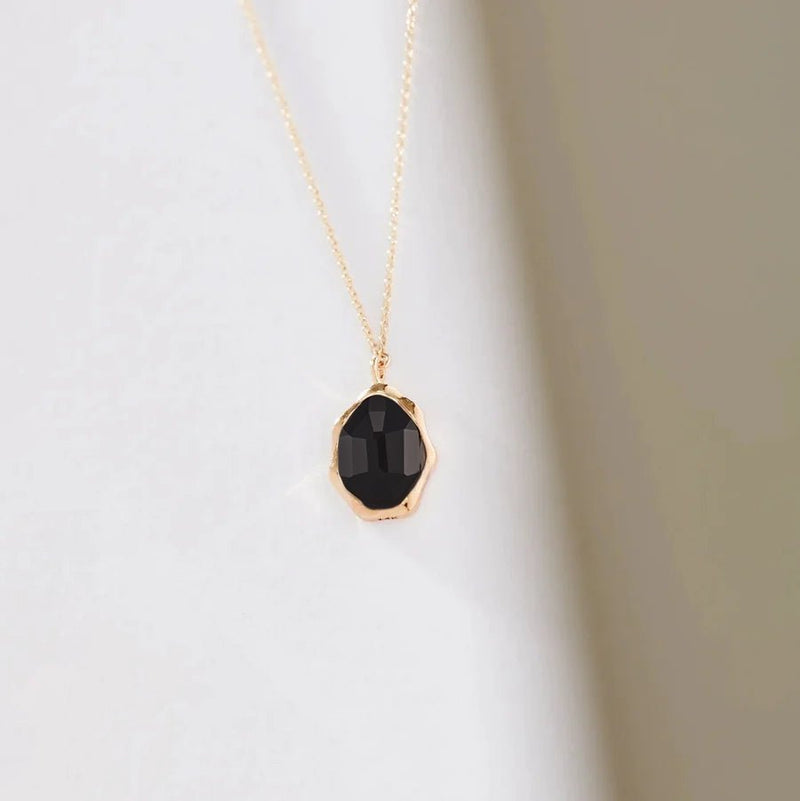 A close-up shot of the bluboho black onyx mood necklace featuring a faceted black gemstone set in a gold frame. The pendant hangs on a delicate gold chain against a plain, light-colored background.