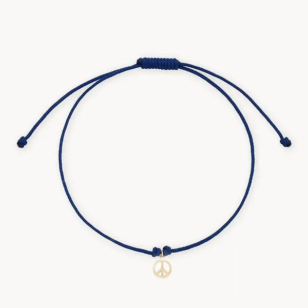 The tie dye moonchild contemplation cord bracelet by bluboho features a minimalist blue cord with an adjustable knot and a small 10k yellow gold peace sign charm hanging from the center. The bracelet’s ends are designed to dangle loosely against a plain white background.